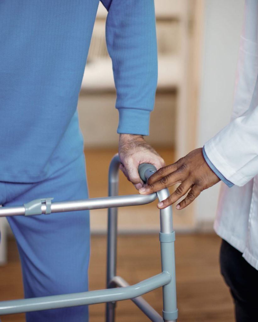 Unrecognizable doctor assisting senior man with mobility walker during physical therapy at nursing home.